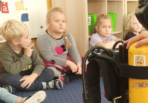 Dzieci siedzą na dywanie i słuchają o pracy strażaka.