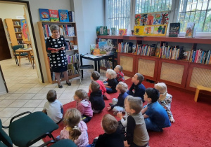 Dzieci siedzą na dywanie w bibliotece i słuchają pani.