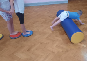 Dzieci stoją na matach sensorycznych, chłopiec toczy się na wałku.