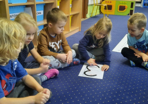 Dzieci siedzą na dywanie w kole, dziewczynka wodzi palcem po konturze cyfry 3.
