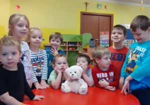 Dzieci pozują do zdjęcia z misiem, który siedzi na stoliku.
