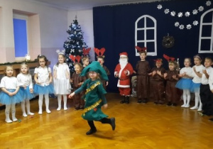 Dzieci ustawione w półkolu śpiewają piosenkę, jedna dziewczynka w przebraniu choinki tańczy na środku sali.