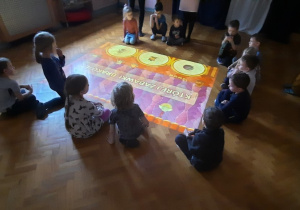 Dzieci siedzą na sali gimnastycznej i obserwują podłogę interaktywną.