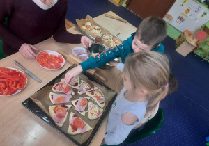 Dzieci siedzą przy stolikach i przygotowują pizzę.