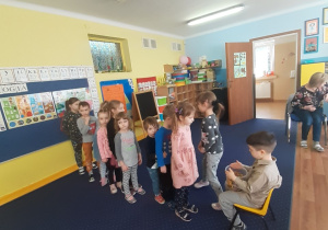 Chłopiec siedzi na krześle, pozostałe dzieci ustawione w rządku składają mu życzenia.