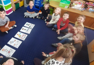 Dzieci siedzą na dywanie w kole, a przed nimi rysunki przedstawiające różne zawody.
