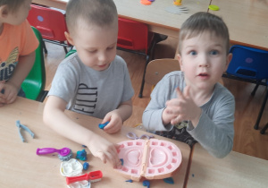 Dzieci siedzą przy stoliku i bawią się prezentując różne zawody.