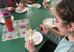 Dzieci siedzą przy stole i nawijają spaghetti na widelec.