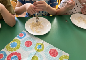 Dzieci siedzą przy stole i nawijają spaghetti na widelec.