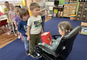 Dzieci ustawione w rzędzie podchodzą do dziewczynki siedzącej na krześle i składają jej życzenia.