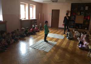 Dzieci siedzą na podłodze, a jeden chłopiec stoi na kartonie z cyframi.