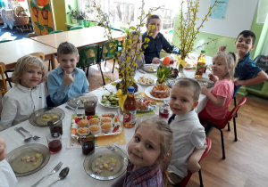 Dzieci siedzą przy stole na którym są smakołyki i biały barszcz.