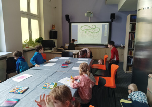 dzieci siedzą przy dużym stole w bibliotece, na którym leżą białe kartony, kredki, w tle na ścianie duża tablica multimedialna