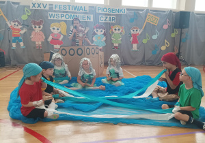 na podłodze niebieski materiał, który imituje wodę, na nim siedzą dzieci, które trzymają wstążki, przypominające fale