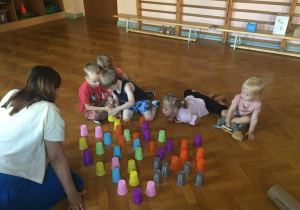 Dzieci siedzą na podłodze i bawią się kolorowymi kubeczkami.
