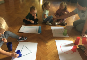 Dzieci siedzą na podłodze i układają kubeczki na figurach geometrycznych.