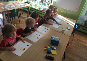 Dzieci siedzą przy stolikach i układają kompozycje z figur geometrycznych.