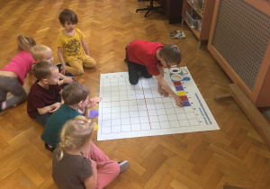 Dzieci siedzą na podłodze i wykonują zadanie na macie do kodowania.