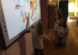 Dzieci siedzą na podłodze i wykonują zadanie na tablicy multimedialnej.