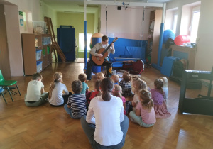 chłopiec gra na gitarze, dzieci siedzą na podłodze na sali gimnastycznej, słuchają muzyki