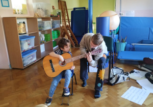 chłopiec trzyma gitarę, starszy chłopiec pokazuje jak prawidłowo chwytać gitarę i na niej grać
