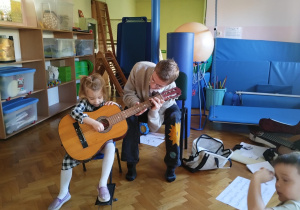 dziewczynka trzyma gitarę, starszy chłopiec pokazuje jak prawidłowo chwytać gitarę i na niej grać