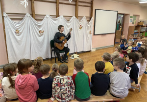 koncert gitarowy na sali gimnastycznej, dzieci siedzą na krzesełkach, chłopiec gra na gitarze