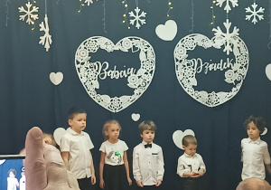 dzieci z grupy 2 występują na sali gimnastycznej, w tle dekoracja z napisami babcia i dziadek