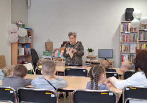wizyta dzieci w bibliotece, dzieci siedzą na krzesełkach przy stolikach, pani bibliotekarka czyta dzieciom książkę
