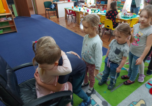 Dziewczynka siedzi na krześle, pozostałe dzieci ustawione w rządku.