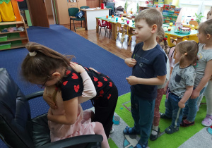 Dziewczynka siedzi na krześle, pozostałe dzieci ustawione w rządku.