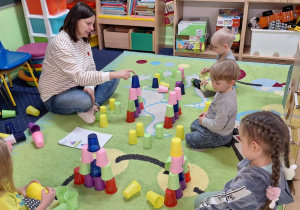 Dzieci siedzą na dywanie i wykonują zadanie z wykorzystaniem kolorowych kubeczków.