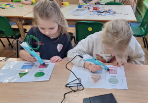 Dzieci siedzą przy stoliku i wykonują zadanie wykorzystując długopis 3D.
