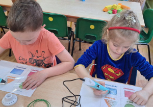 Dzieci siedzą przy stoliku i wykonują zadanie wykorzystując długopis 3D.