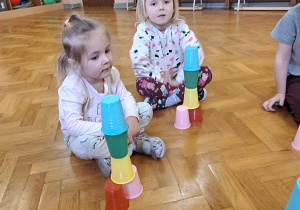 Dzieci siedzą na podłodze i układają kolorowe kubeczki.