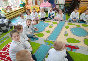 Dzieci siedzą w kole w białych fartuszkach laboratoryjnych.