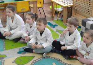 Dzieci siedzą w kole w białych fartuszkach laboratoryjnych.