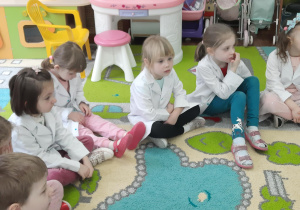 Dzieci siedzą w kole w białych fartuszkach laboratoryjnych.