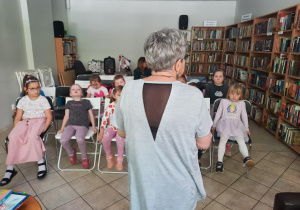 Dzieci siedzą na krzesłach i słuchają pani bibliotekarki.
