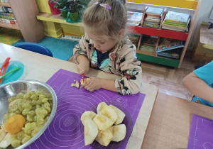 dziewczynka siedzi przy stoliku i kroi owoce na sałatkę owocową.