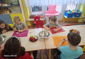 dzieci siedzą przy stolikach, kroją owoce i warzywa na kolorowych matach