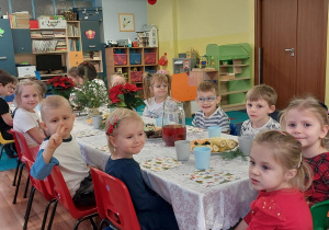 dzieci w odświętnych strojach siedzą przy stolikach, na których widać talerze ze smakołykami, ciastami, babeczkami.