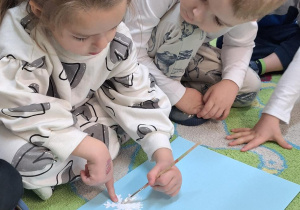 Dzieci siedzą na dywanie i wykonują pracę plastyczną.