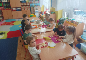 dzieci siedzą przy stolikach, kroją owoce na sałatkę