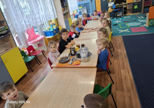 dzieci siedzą przy stolikach, na tacy stoją produkty spożywcze potrzebna do zrobienia słodyczy