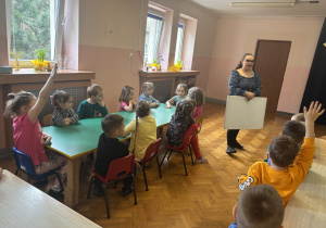 Dzieci siedzą przy stolikach i odpowiadają na pytania.