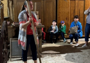 Dzieci siedzą w stodole i oglądają sprzęty wykorzystywane dawniej na wsi.