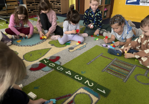 Dzieci siedzą na dywanie i bawią się klockami Lego duplo oraz poznają cyfry.