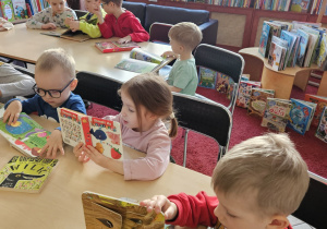 Dzieci siedzą przy stolikach i oglądają książki.
