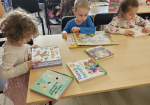 Dzieci siedzą przy stolikach i oglądają książki.